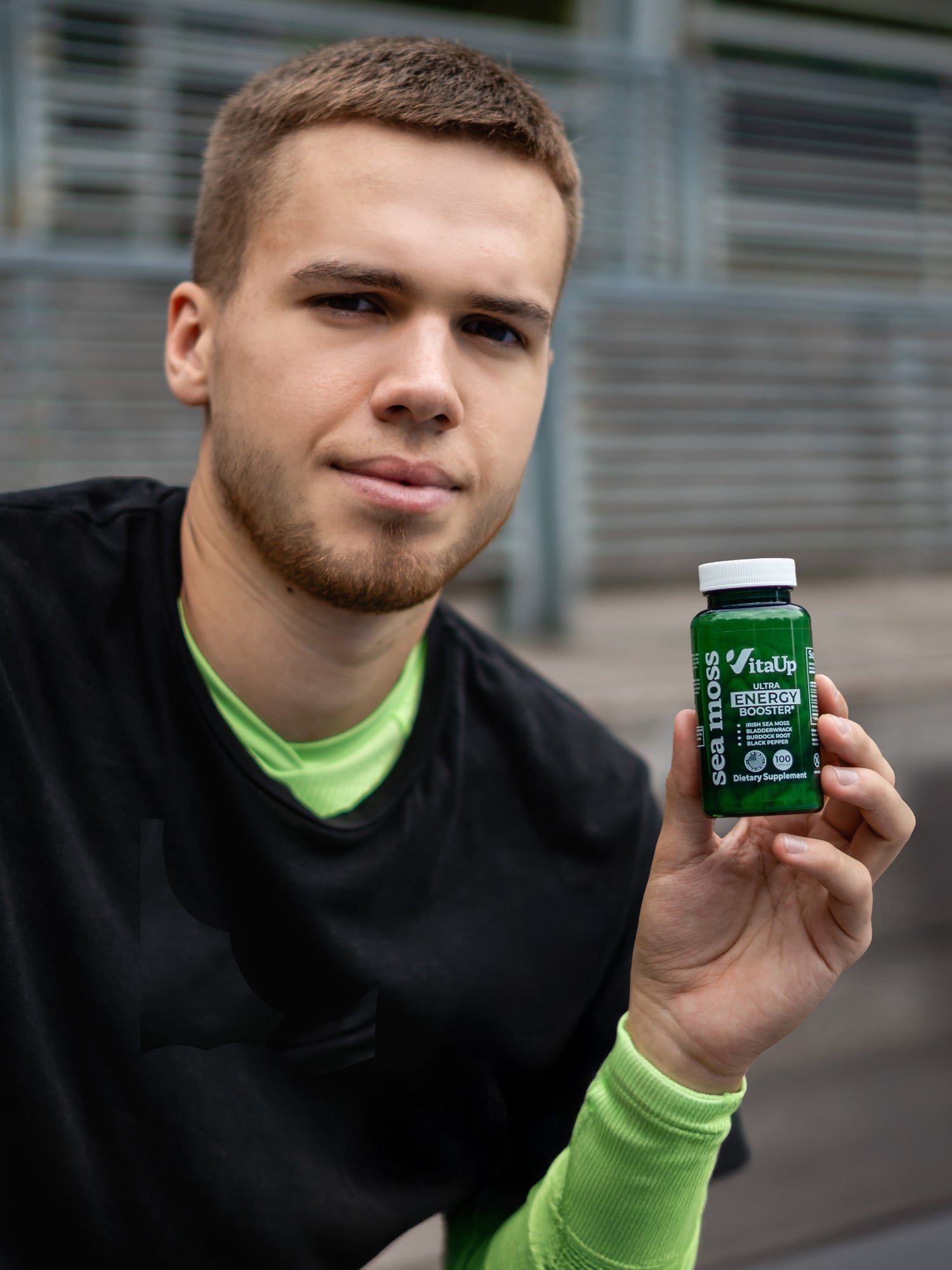 Smiling young man holding a VitaUp Sea Moss supplement bottle, showcasing product with confidence outdoors.