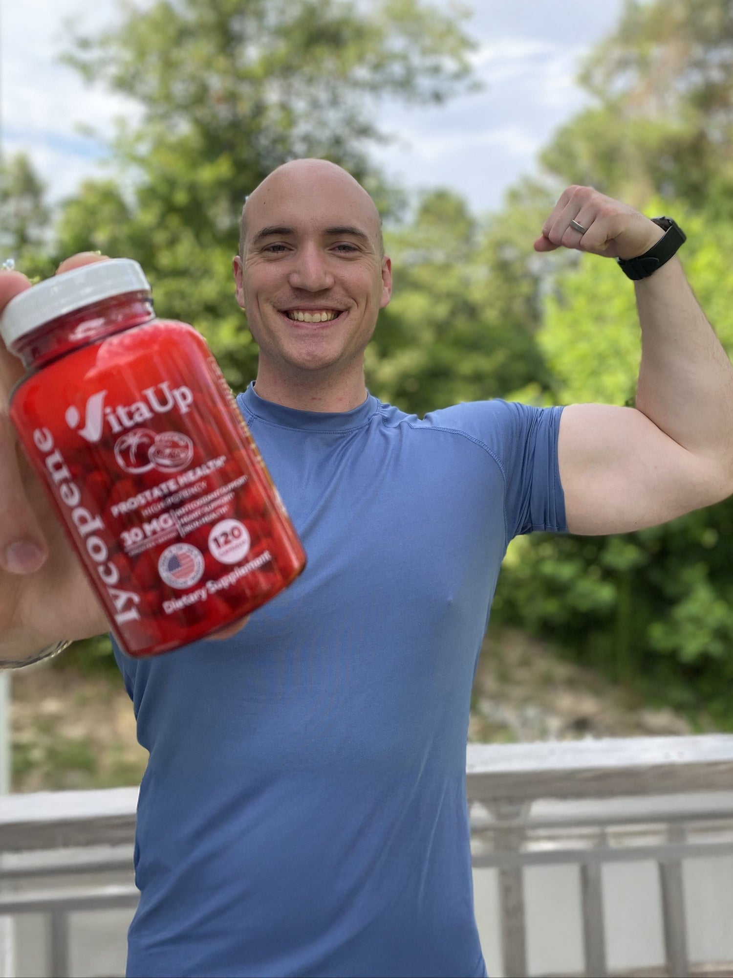 Smiling man outdoors flexing biceps while holding VitaUp Lycopene supplement bottle, showcasing energy and fitness benefits.
